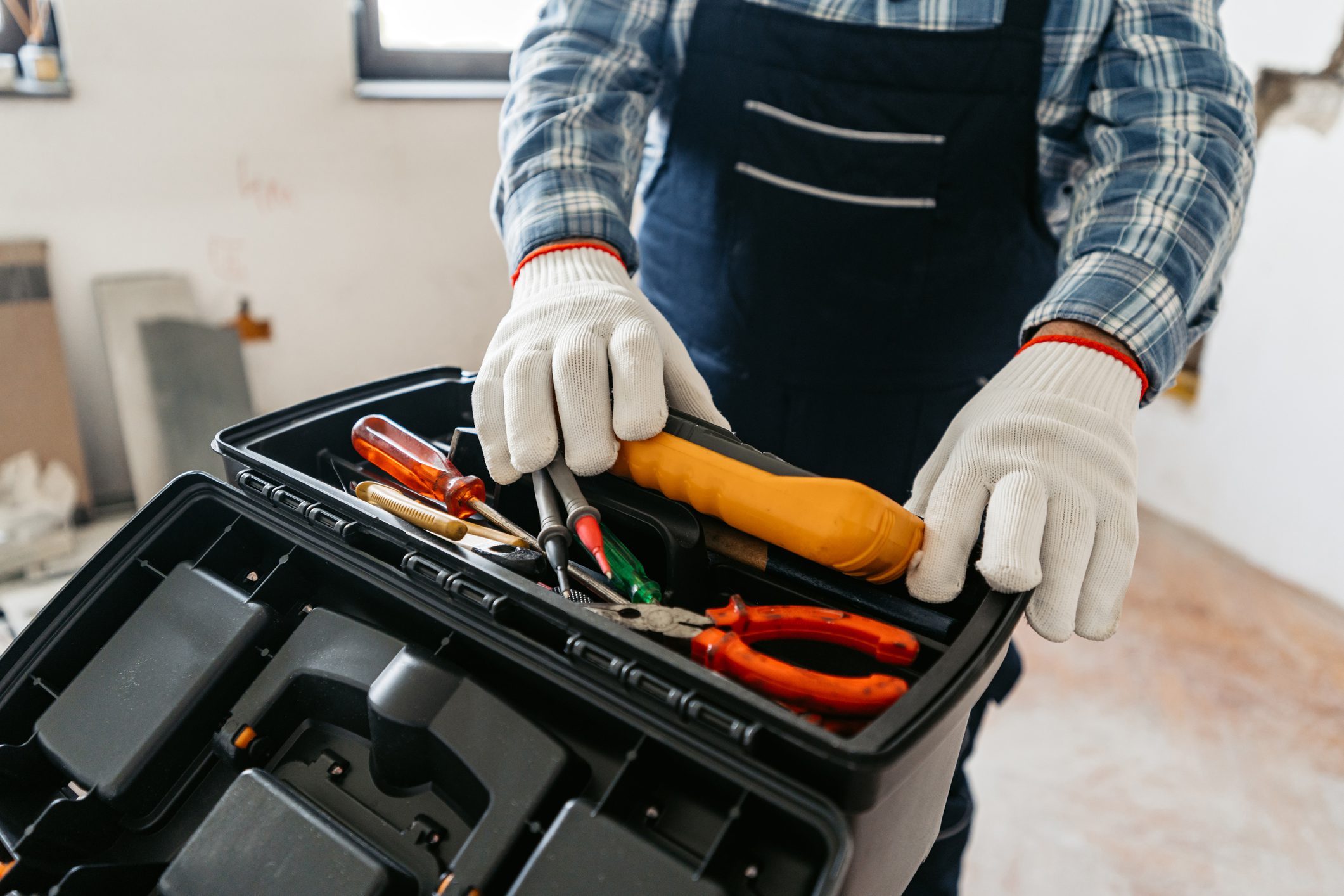 Senior male electrician in overalls putting a multimeter in a tool box in an empty apartment under renovation. Close-up.